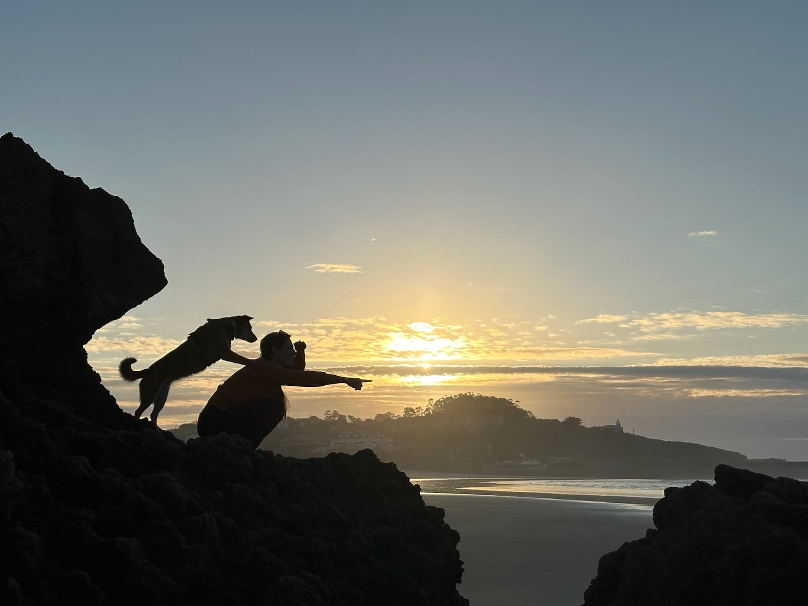 Laure kauert bei Sonnenaufgang auf Felsen und zeigt zum Horizont. Lina hat die Pfoten auf ihrer Schulter und schaut in dieselbe Richtung.